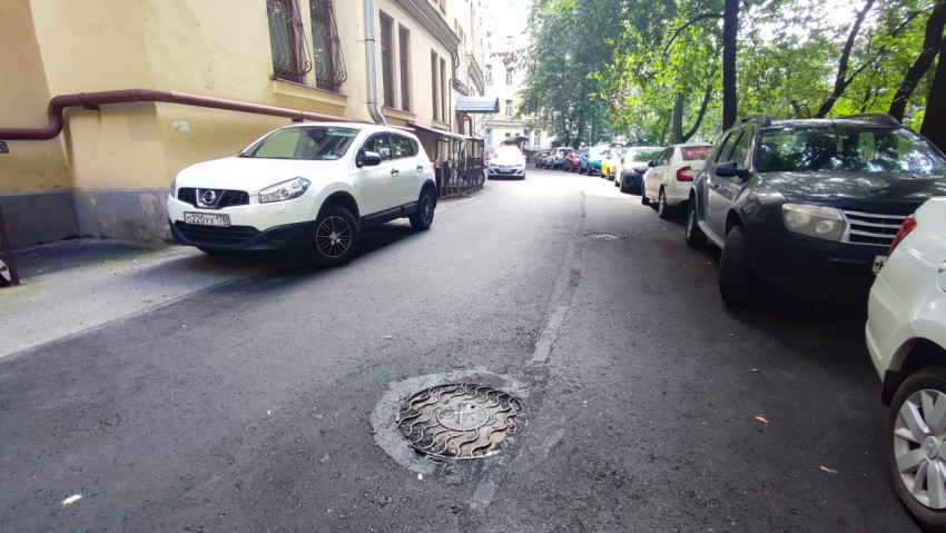 Свежий асфальт во дворовом проезде с люком канализации и припаркованными машинами вдоль фасада