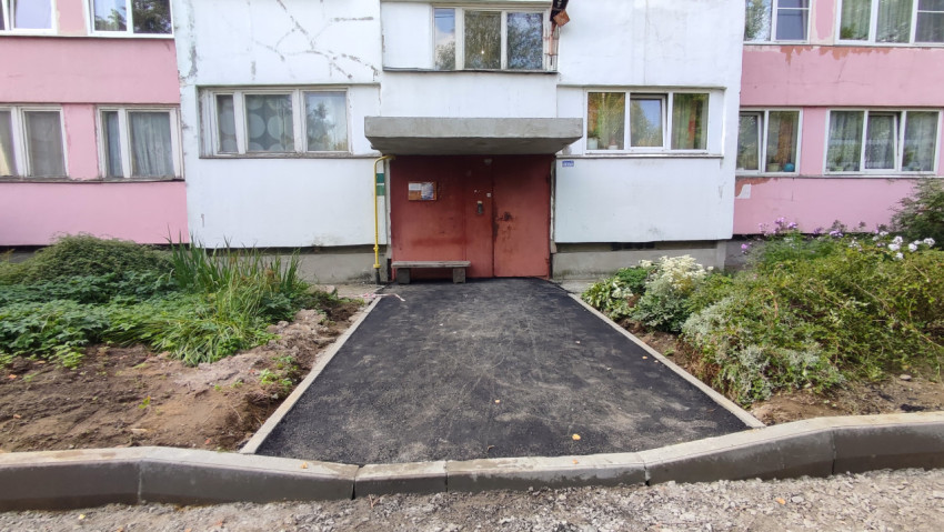 Новый асфальтовый подход к подъезду с красной дверью проходит между клумбами и бордюрами