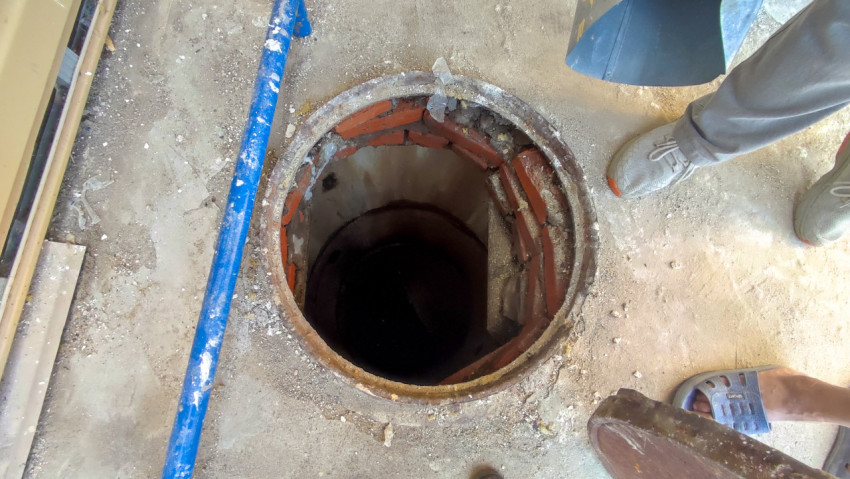 Открытый технический люк в бетонном полу с кирпичной шахтой и инструментом при обслуживании коммуникаций