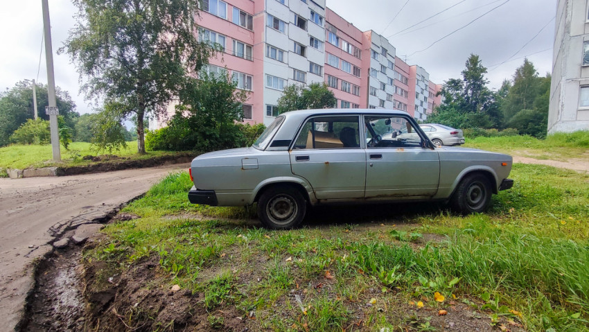 Профильный вид седана Lada на траве у повреждённого края дороги между домами