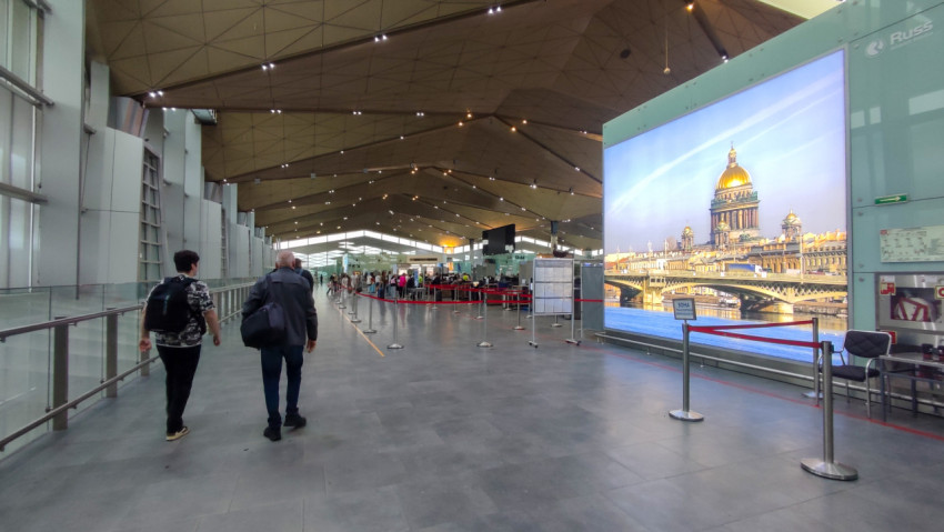 Внутри терминала Пулково люди идут по просторному залу мимо большого экрана