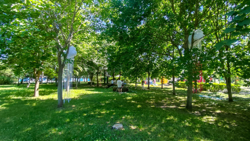 Тенистая поляна в городском парке с подвесными арт-объектами на деревьях и рабочими на газоне