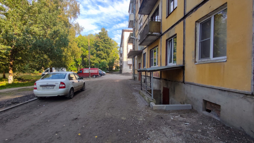 Грунтовый дворовой проезд вдоль жёлтого дома с припаркованными машинами и зелёными деревьями