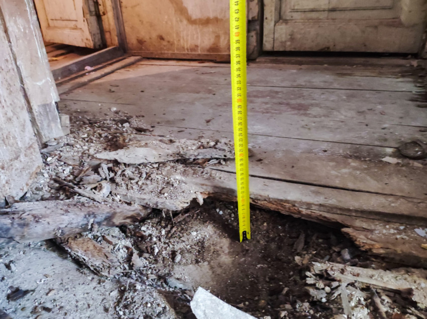 Замер глубины провала в деревянном полу рулеткой у дверного проёма