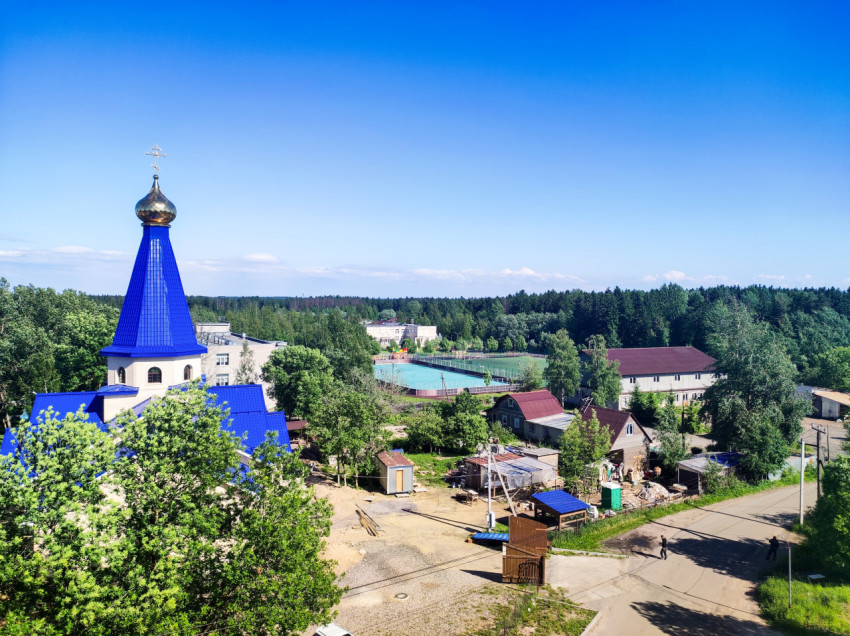 Православный храм с синим шатром и золотым куполом на фоне посёлка и леса
