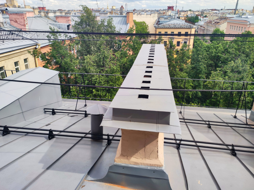 Кровельный вентиляционный короб на кирпичном основании на фоне городских крыш и деревьев