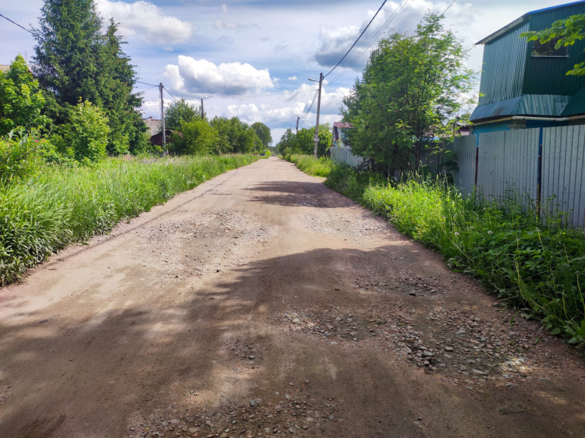 Грунтовая улица с гравийными участками, металлическим забором и придорожной травой