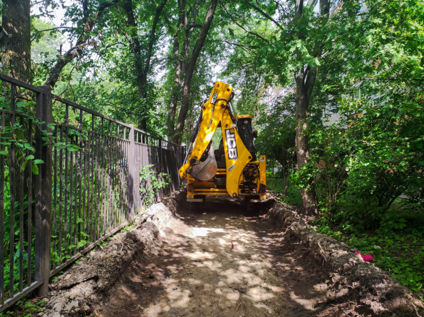 Экскаватор-погрузчик JCB на разрытом проезде рядом с ограждением и деревьями