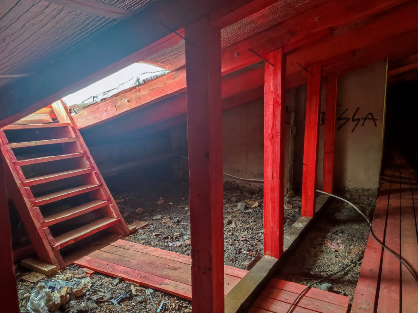 Чердачное помещение с красными подпорками, деревянной лестницей к проёму и строительным мусором на полу