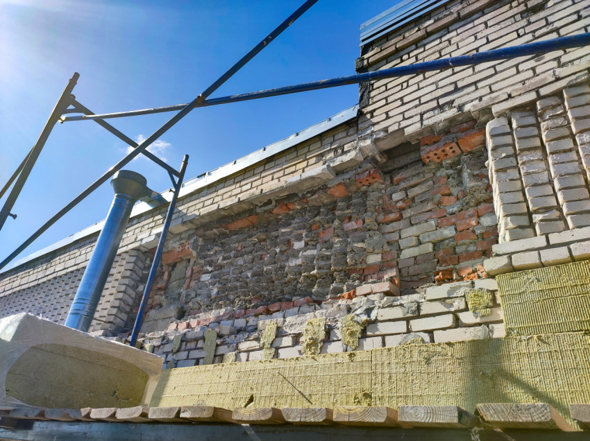 Участок фасада с очищенной кирпичной стеной, водосточной трубой и полосой утеплителя на лесах