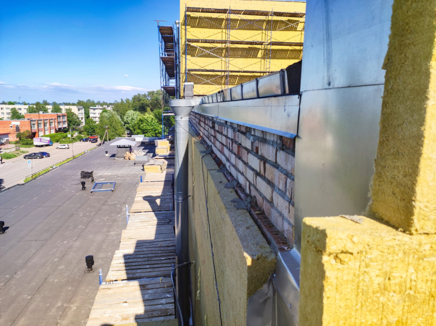 Вид вдоль парапета с утеплителем, отливами и водосточной трубой на крыше, на фоне городской улицы и лесов