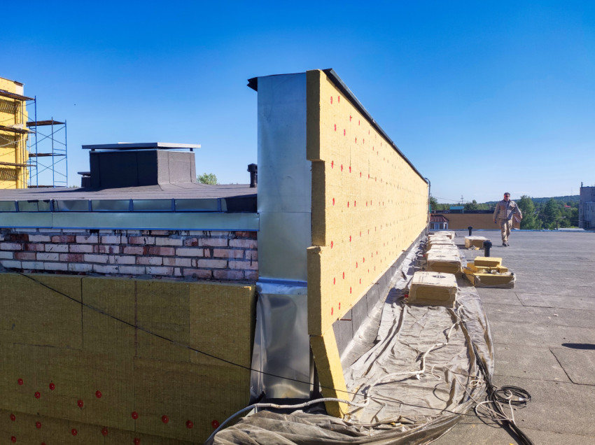 Рабочий на плоской крыше рядом с утепляемым парапетом и строительными материалами