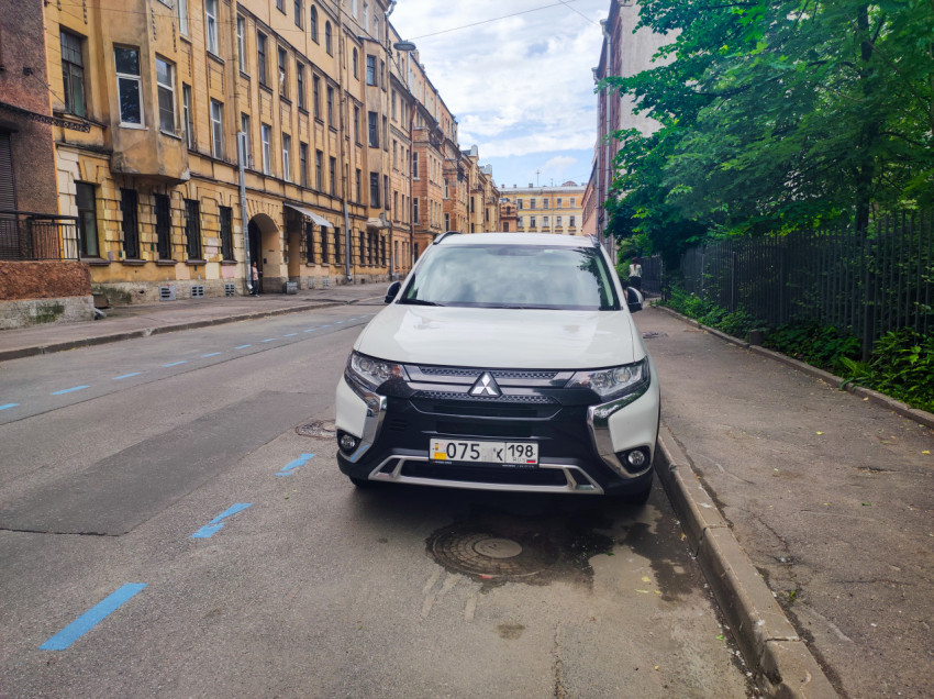 Белый кроссовер Mitsubishi Outlander припаркован у тротуара на городской улице