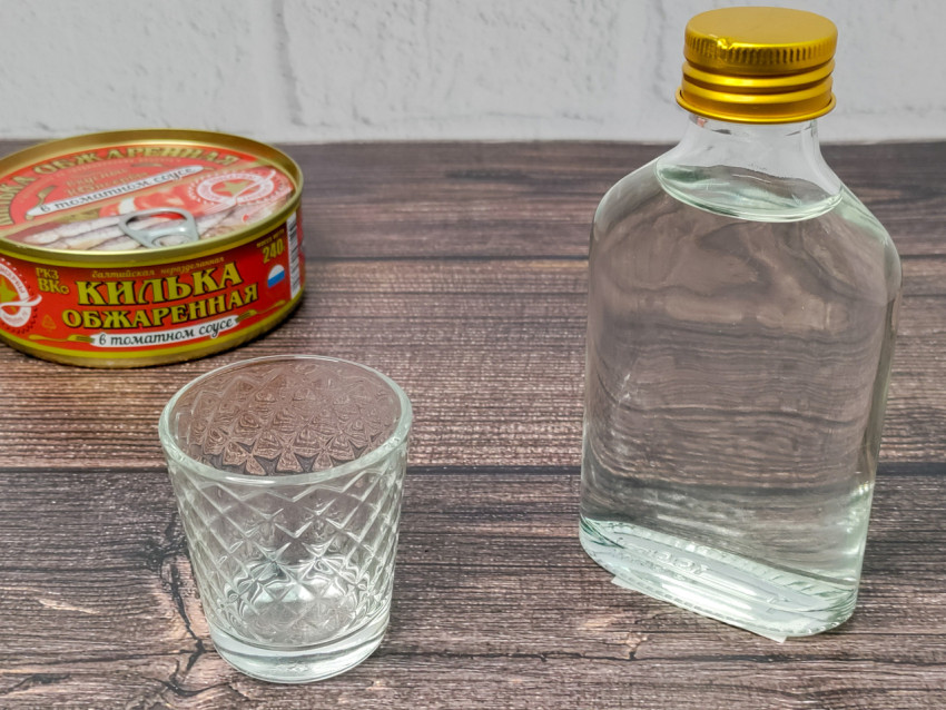 Стеклянная рюмка, бутылка и банка кильки в томатном соусе на деревянном столе