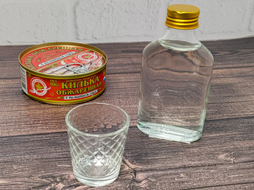 Консервы кильки, бутылка и рюмка на деревянном столе