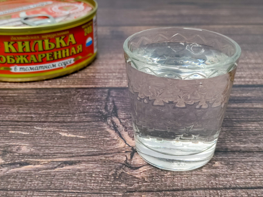 Рюмка водки и банка кильки на деревянной поверхности