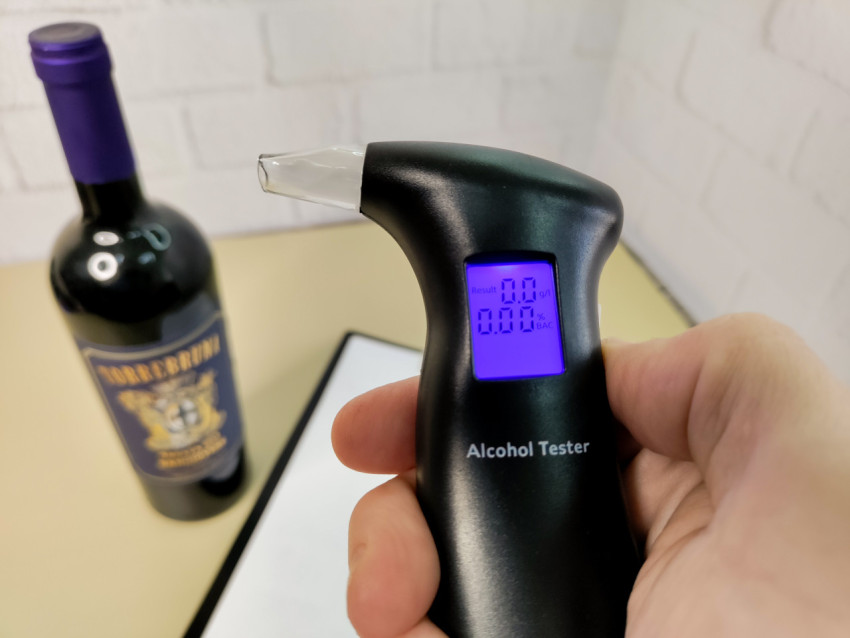Рука держит алкотестер с нулевым результатом перед бутылкой вина
