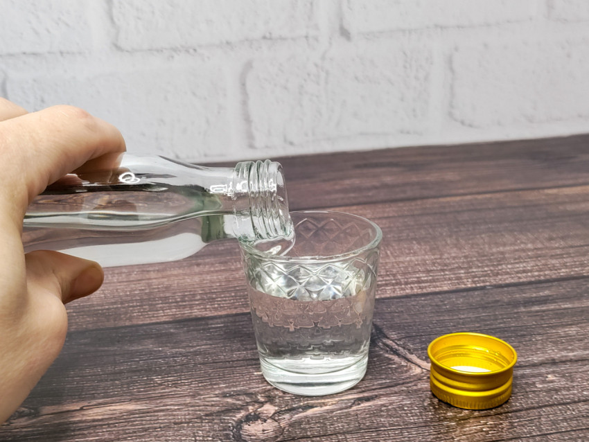 Рука наливает водку в рюмку, стоящую на деревянной поверхности