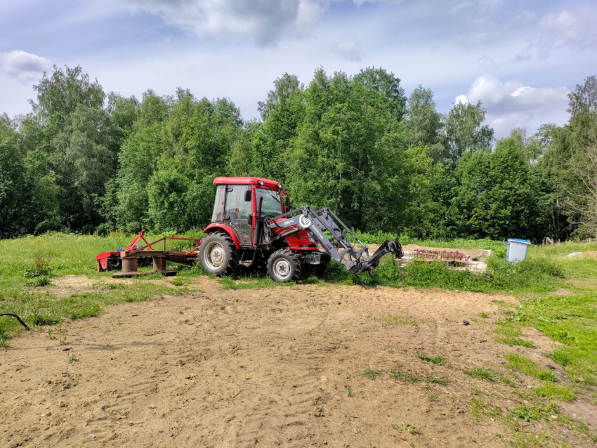 Красный трактор с фронтальным погрузчиком на грунтовой площадке у леса