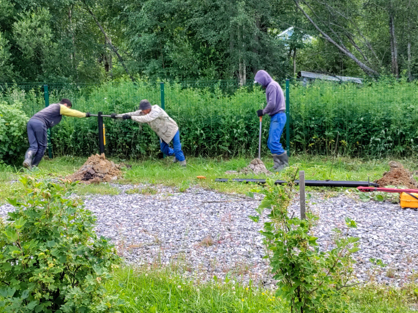 Рабочие вручную завинчивают винтовую сваю для свайного фундамента на участке у забора