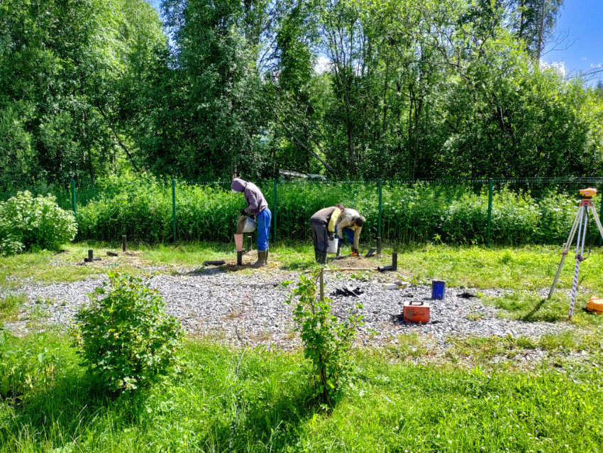 Рабочие заполняют металлические сваи смесью, рядом установлен геодезический нивелир