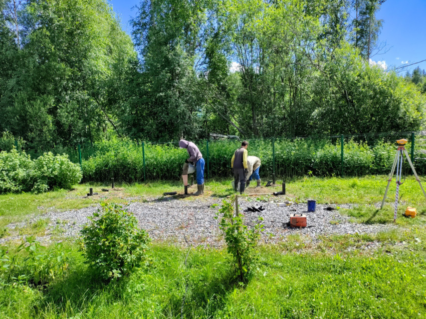 Монтаж свайного фундамента: рабочие с вёдрами и геодезический прибор на площадке у ограждения
