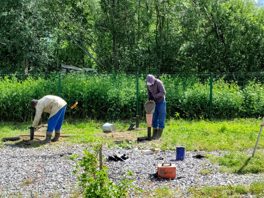 Рабочие подсыпают и выравнивают грунт вокруг винтовых свай при монтаже свайного фундамента