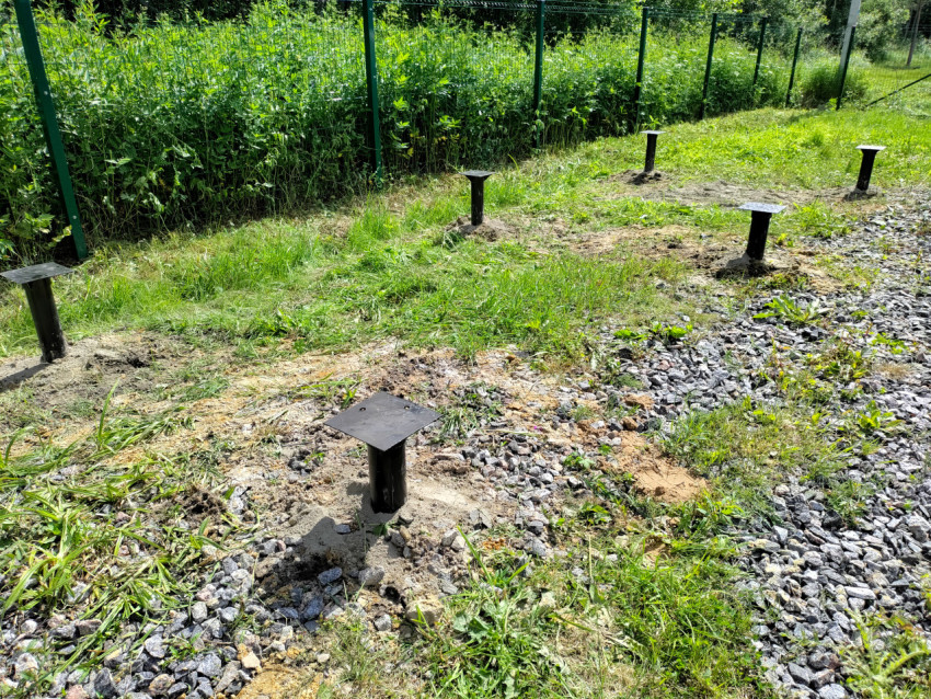 Ряд установленных винтовых свай свайного фундамента на травяной площадке вдоль ограждения