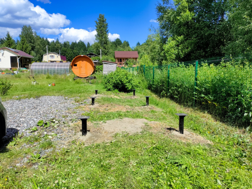 Свайный фундамент под дом на участке на фоне теплицы, домов и бани-бочки