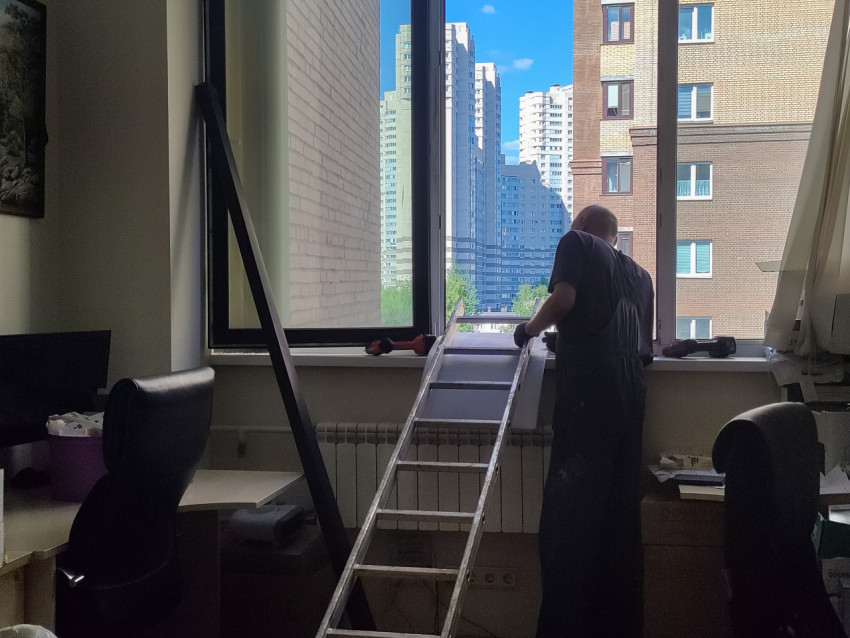 Рабочий устанавливает элементы у офисного окна с видом на жилые высотки