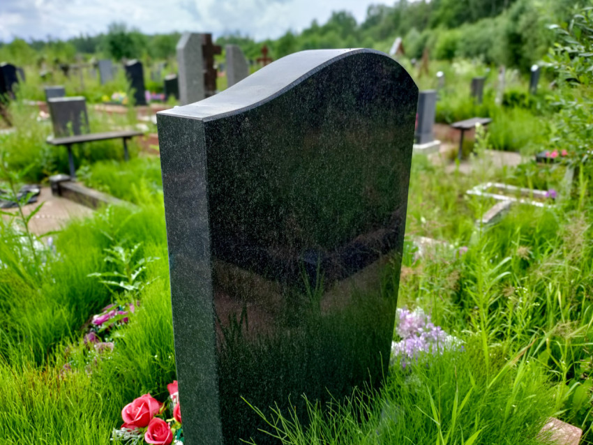 Полированная гранитная стела на кладбище среди высокой травы и цветов