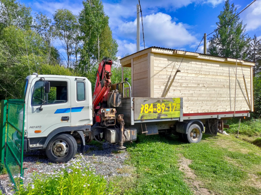 Грузовик с краном-манипулятором перевозит деревянную бытовку по загородной дороге