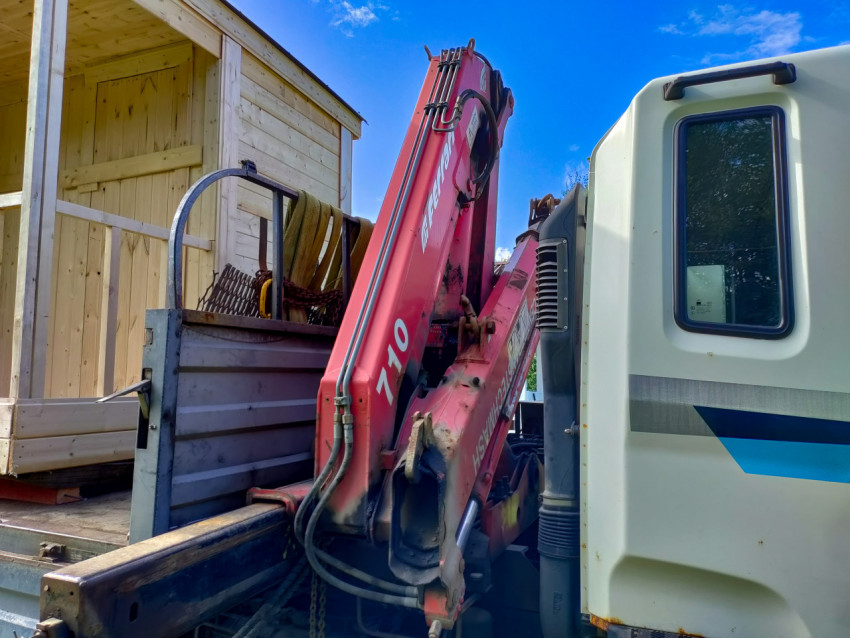 Стрела крана-манипулятора с гидрошлангами на грузовике рядом с деревянной бытовкой перед разгрузкой