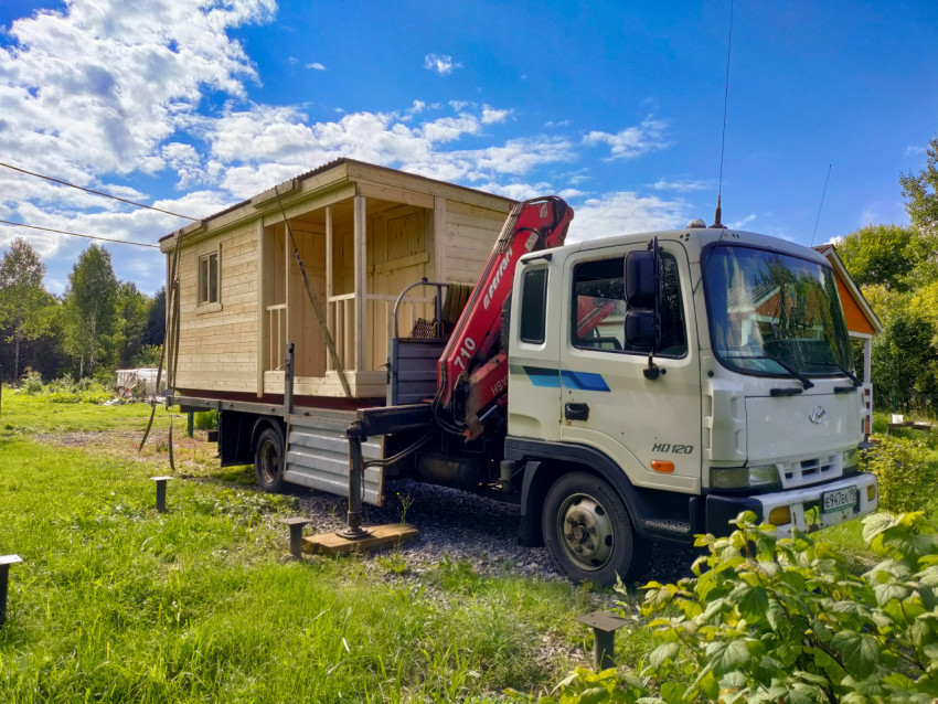 Грузовик с краном-манипулятором и деревянной бытовкой на дачном участке среди травы и деревьев