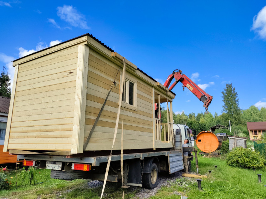 Кран-манипулятор разгружает деревянную бытовку на дачном участке, рядом стоит рабочий и видны винтовые сваи