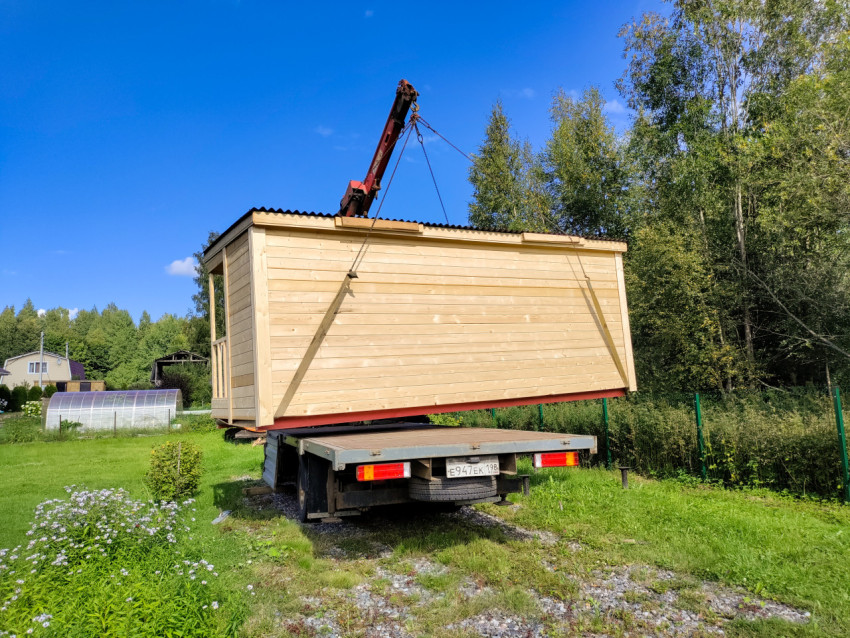 Кран-манипулятор переносит деревянный модульный дом над дачным участком рядом с теплицей