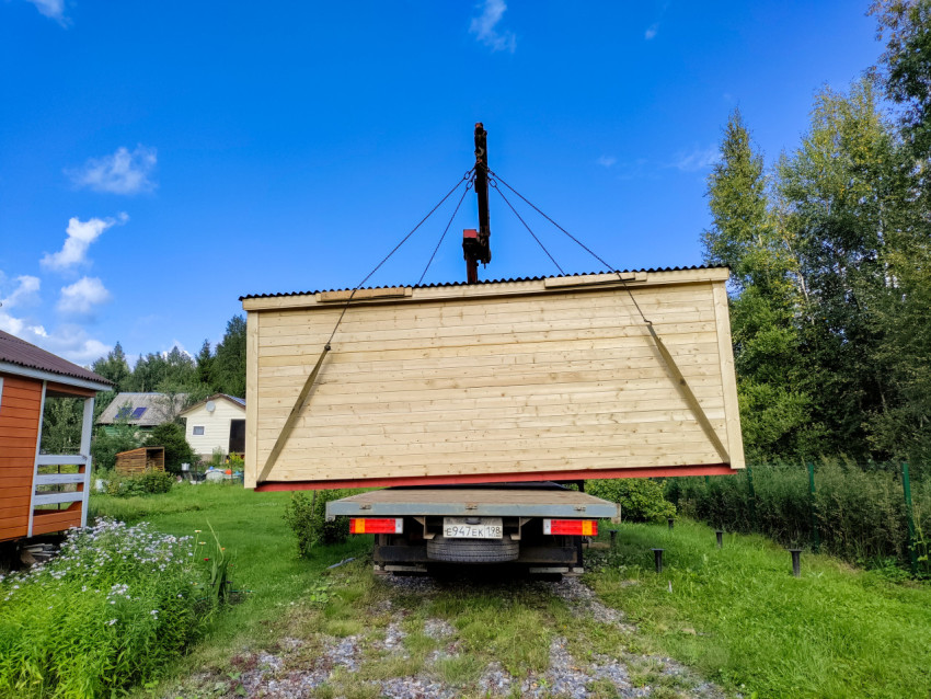 Деревянный модульный дом подвешен на стропах за краном-манипулятором перед установкой на участке