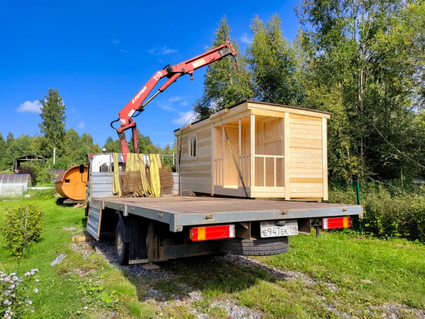 Кран-манипулятор переносит деревянную бытовку над участком с зелёными деревьями