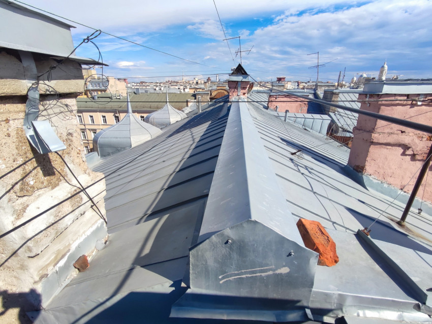 Вид по коньку фальцевой крыши с куполами, дымоходами, антеннами и натянутыми проводами над городом