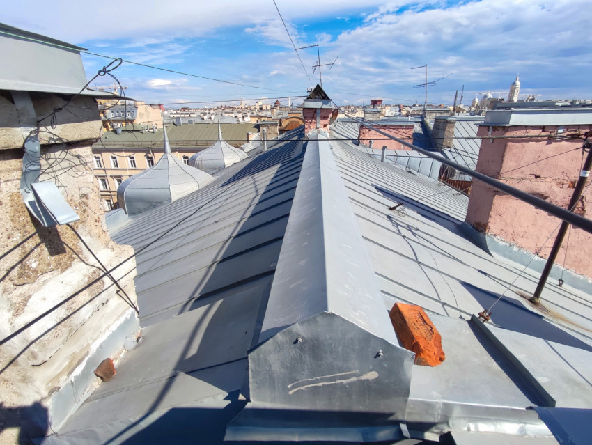 Фальцевая крыша с коньком, дымоходами и кабелями молниезащиты на фоне городской панорамы и облаков