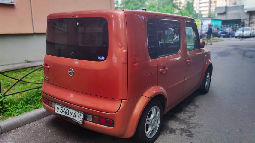 Задний левый ракурс Nissan Cube на парковке с видимыми вмятинами