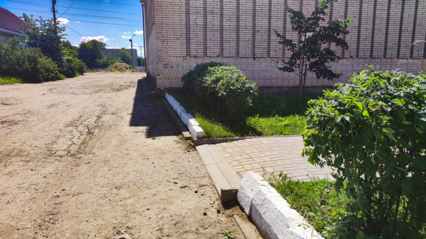 Разбитая грунтовая дорога у здания с тротуаром, бордюром и зарослями кустов