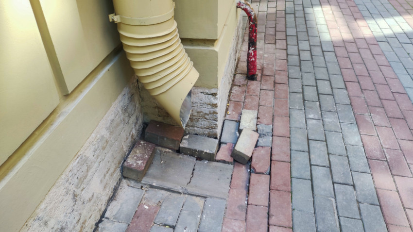 Водосточный слив у фасада и повреждённая тротуарная плитка у цоколя
