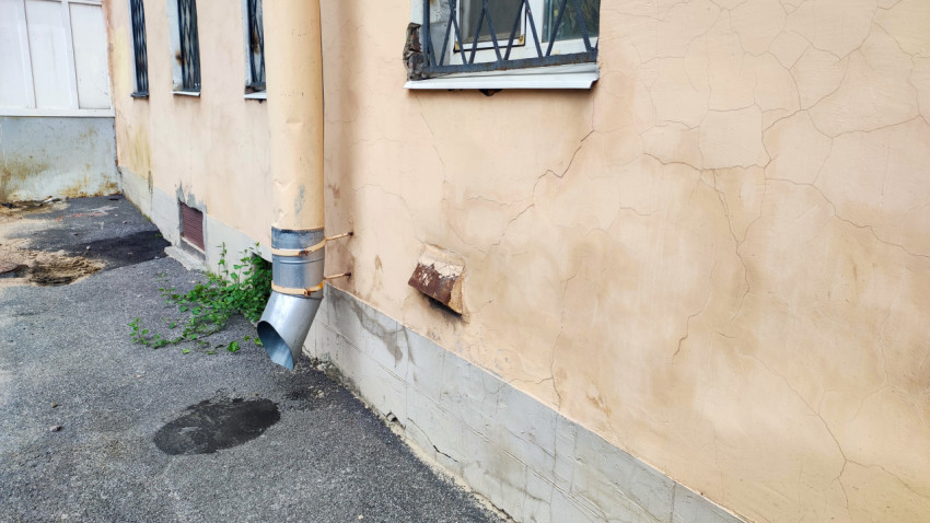 Угол фасада с водосточной трубой и заметной трещиной у основания здания рядом с отмосткой