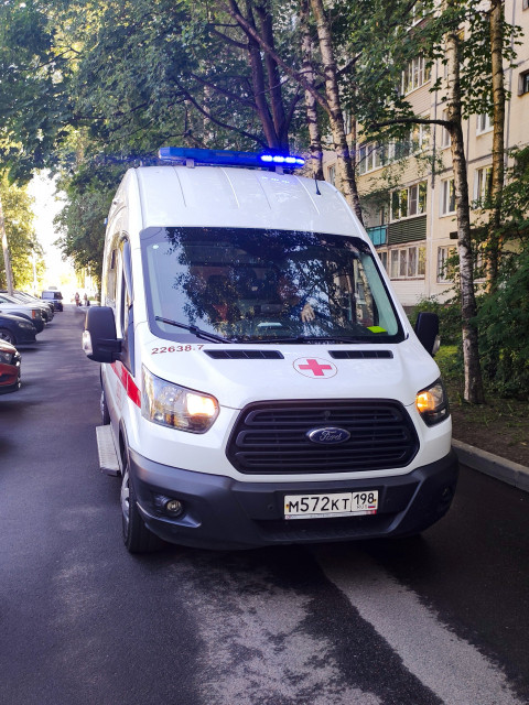 Передний план автомобиля скорой помощи Ford Transit с включёнными маячками на дворовой дороге