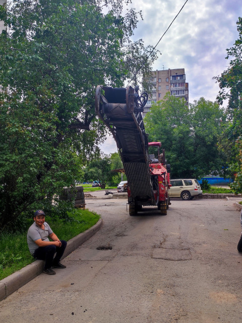Дорожная фреза с поднятым конвейером стоит во дворе, рядом сидит рабочий на бордюре