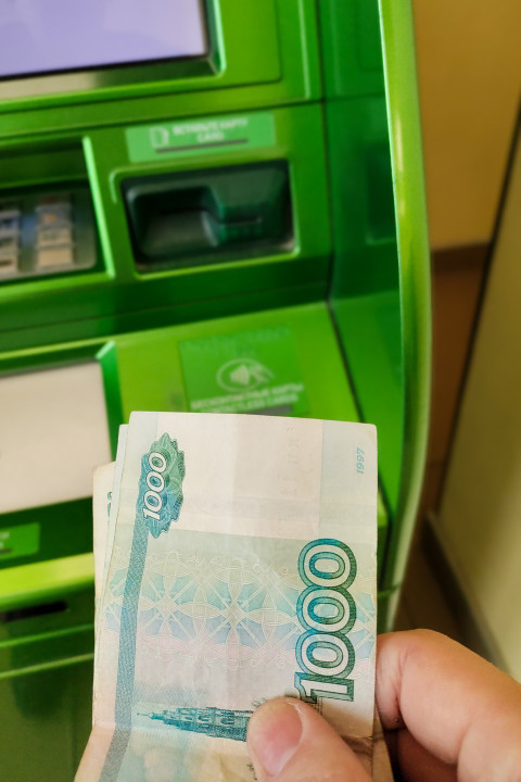 Рука держит купюры перед банкоматом Сбербанка