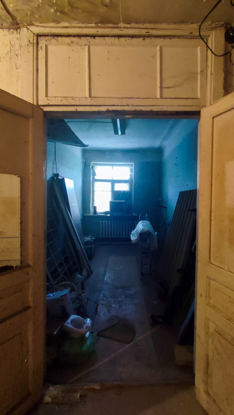 Вид через старые двустворчатые двери в пустующее помещение с окном, радиатором и строительными материалами