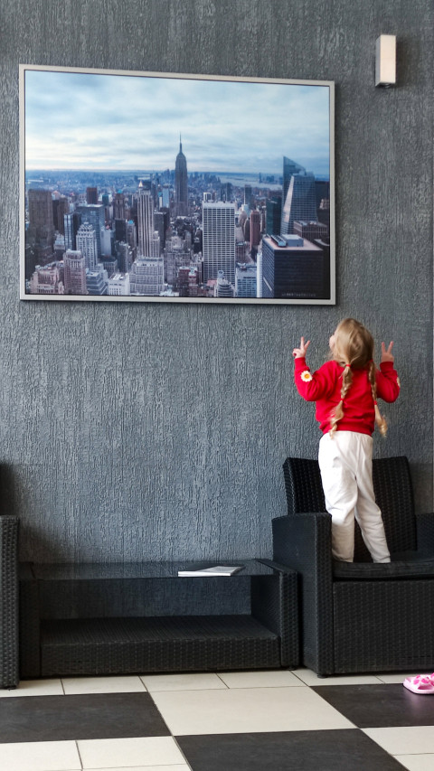 Девочка с косичками стоит на кресле и показывает жесты перед постером города