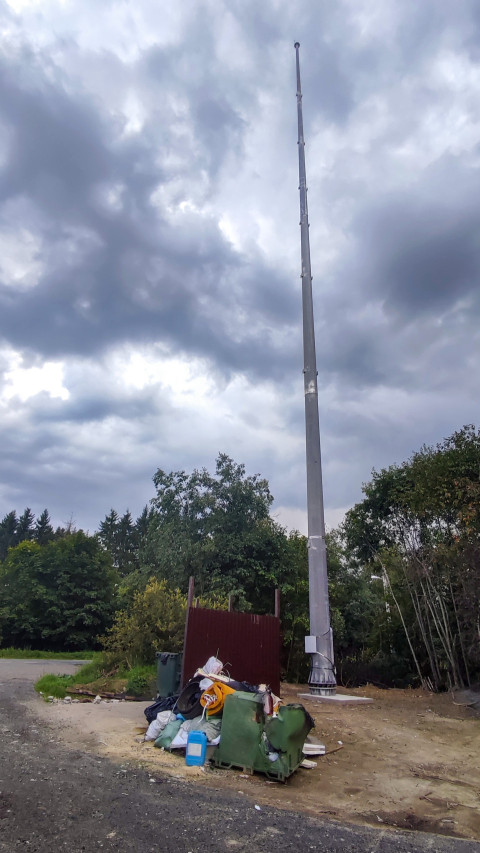 Высокая металлическая мачта освещения на фоне облачного неба и деревьев у технической площадки